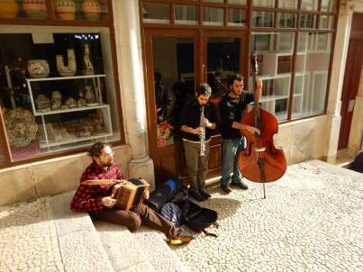 three musicians play on the streets of Portugal. One man in a checkered shirt plays the accordian on the floor. The middle man plays a brass instrument and the final man is playing bass.