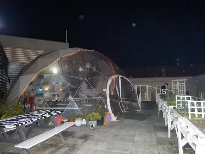 a plastic dome tent with a bar and seating inside in Lisbon Portugal on a roof top. It is night time and raining