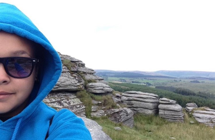 Close Up shot of a girl in a bright blue hoodie on the cliffs of dartmoor with the rocks and lush green hills rolling behind her
