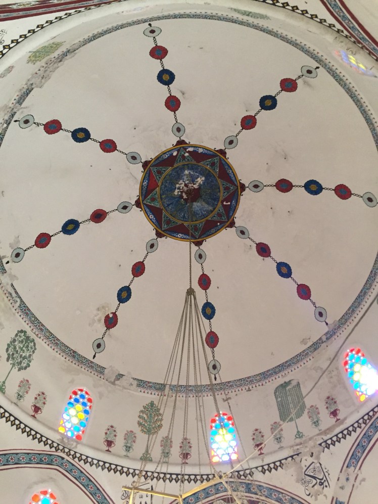 the ceiling of a mosque. There is a star like pattern in the centre. From each point you can see lines of bead like shapes are coming from it towards the edges of the ceiling. Small windows below this reveal brightly coloured light from the sun through the stain-glass windows