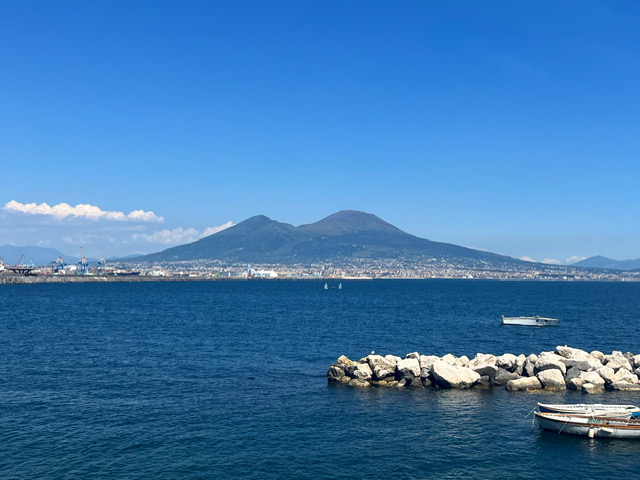 The sea takes up most of the image. In the centre back Mount Vesuvius sits beneath clear blue sky. 