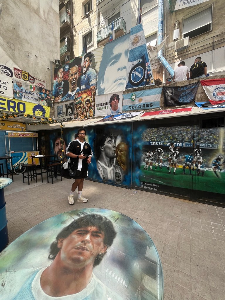 A man stands in a football shirt surrounded by images of maradonna on the walls and floor. 